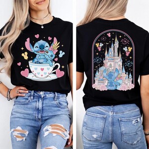May include: Black t-shirt featuring a cartoon Stitch character in a teacup with a friend, surrounded by hearts and stars. The back of the shirt shows Stitch in front of a castle with fireworks and flowers. The design is in shades of blue, pink, and yellow.