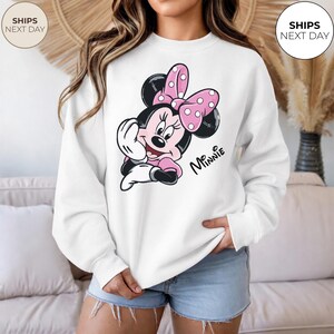May include: White sweatshirt featuring a Minnie Mouse graphic. The graphic shows Minnie Mouse in a pink dress and bow with white polka dots. The word "Minnie" is written in cursive. The sweatshirt is a crew neck style.