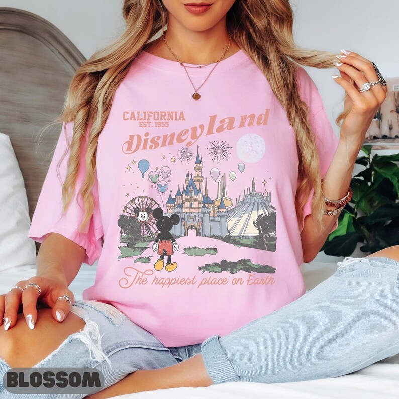 May include: Pink t-shirt featuring a Disneyland graphic. The design includes a castle, Mickey Mouse, and the text "California Disneyland Est. 1955" and "The happiest place on Earth".