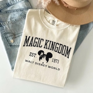 May include: Cream-colored t-shirt with the text "MAGIC KINGDOM" in black, arched across the top. Below, it reads "EST. 1971 WALT DISNEY WORLD" with a Mickey Mouse silhouette. The shirt is paired with light-wash jeans and a tan hat.