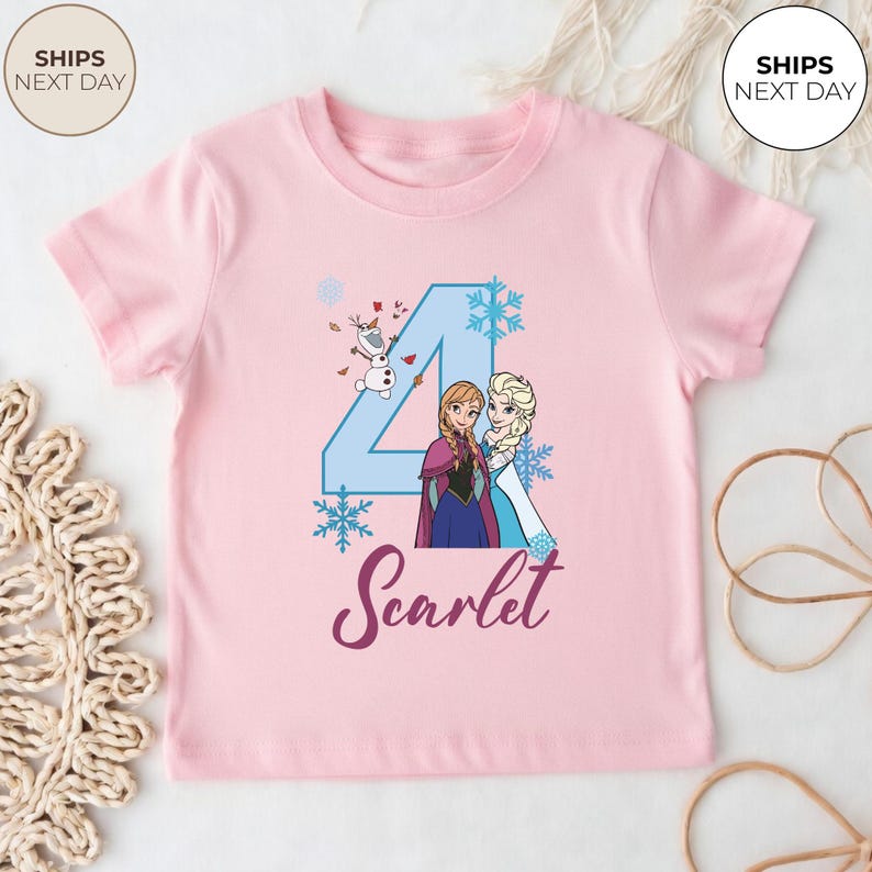 May include: A pale pink t-shirt with a blue number 4, snowflakes, and the Frozen characters Anna and Elsa. The name "Scarlet" is written in a cursive font below the number.
