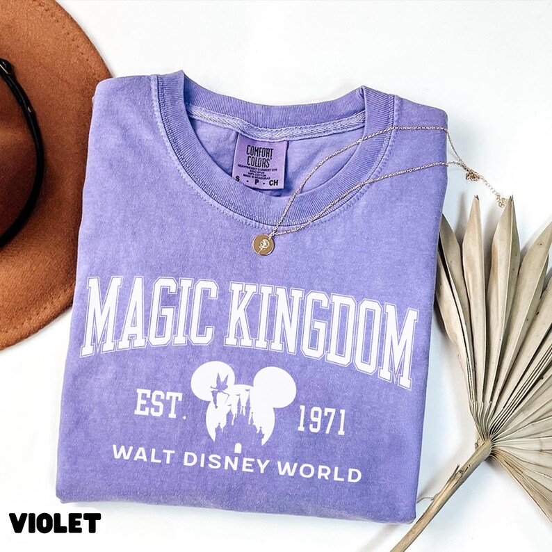May include: A violet Comfort Colors t-shirt with the words "MAGIC KINGDOM" in white block letters. Below the text is the silhouette of Mickey Mouse ears and a castle, with "EST. 1971" and "WALT DISNEY WORLD" printed below. A gold necklace rests on the shirt.
