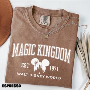 May include: Espresso-colored t-shirt with "MAGIC KINGDOM" printed in white block letters. Below the text is a graphic of the Disney castle and Mickey Mouse ears, with "EST. 1971" and "WALT DISNEY WORLD". A silver necklace with a pendant rests on the shirt.