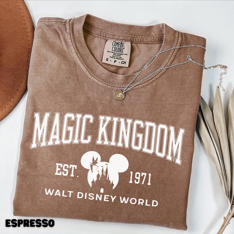 May include: Espresso-colored t-shirt with "MAGIC KINGDOM" printed in white block letters. Below the text is a graphic of the Disney castle and Mickey Mouse ears, with "EST. 1971" and "WALT DISNEY WORLD". A silver necklace with a pendant rests on the shirt.