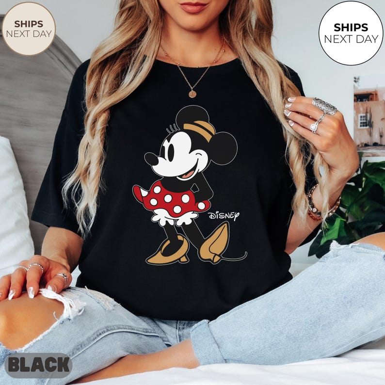 Puede incluir: Camiseta negra con un dibujo de Minnie Mouse. El personaje de dibujos animados lleva una falda roja de lunares blancos, zapatos amarillos y un sombrero amarillo y negro. La palabra "Disney" est&aacute; impresa debajo del personaje.