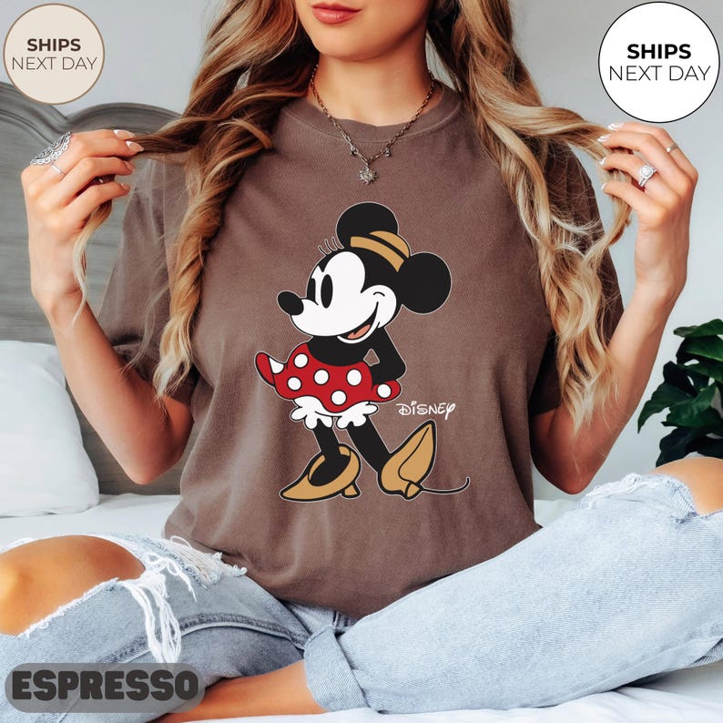 Puede incluir: Camiseta color espresso con un gr&aacute;fico de Minnie Mouse. El personaje de dibujos animados lleva un vestido de lunares rojos y blancos, zapatos amarillos y un lazo negro y amarillo. El logotipo de Disney est&aacute; impreso debajo.