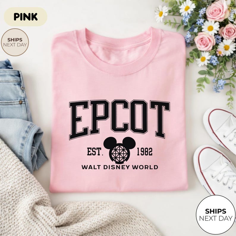 May include: A light pink t-shirt with the word "EPCOT" in black block letters. Below the word "EPCOT" is the text "EST. 1982 WALT DISNEY WORLD" in black. A Mickey Mouse head graphic is between "EST." and "1982".