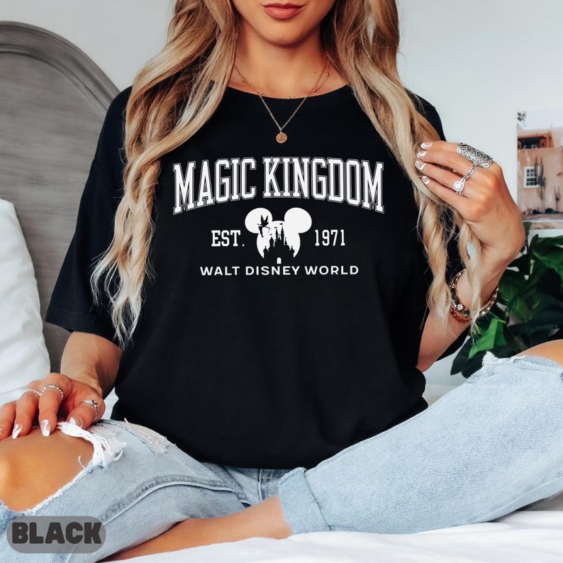 May include: Black t-shirt with the text "MAGIC KINGDOM EST. 1971 WALT DISNEY WORLD" in white. The design includes a silhouette of a castle and Mickey Mouse ears. The shirt is worn with blue jeans.