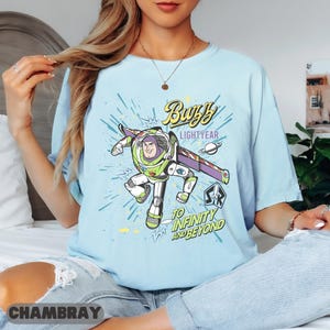 Może przedstawiać: Jasnoniebieska koszulka z grafiką Buzza Astrala z Toy Story. Projekt zawiera tekst "Buzz Lightyear" i "To Infinity and Beyond" w stylu komiksowym. Koszulka jest wykonana z miękkiego materiału.