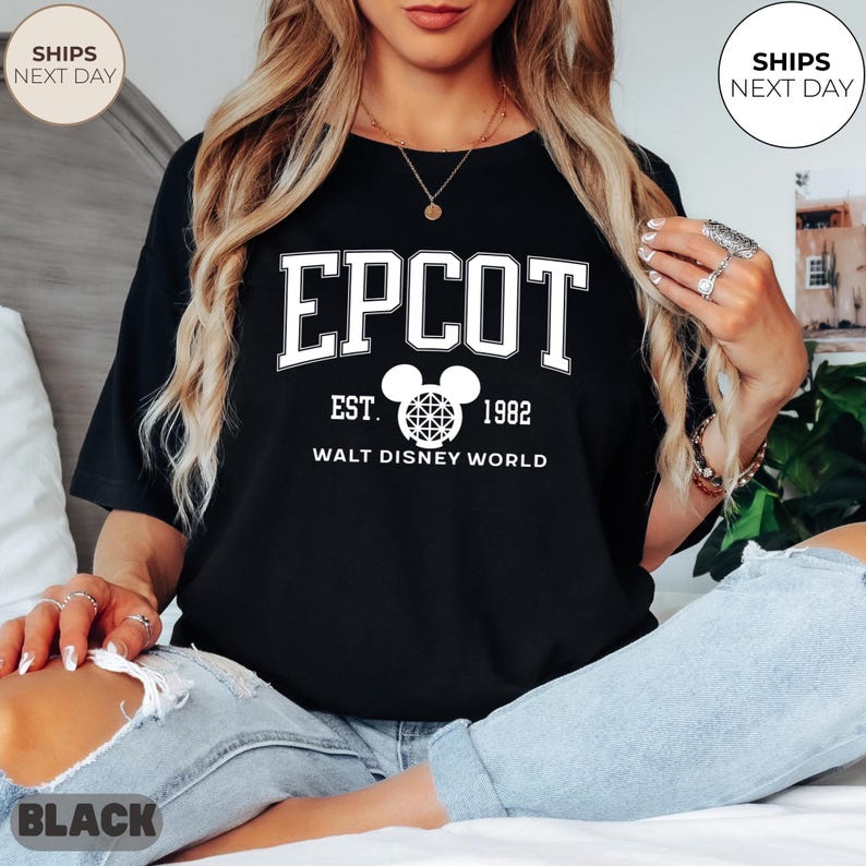 May include: Black t-shirt with the word "EPCOT" in white block letters, with "EST. 1982" and "WALT DISNEY WORLD" below. A Mickey Mouse head graphic is in the center. The shirt is worn by a person wearing blue jeans.