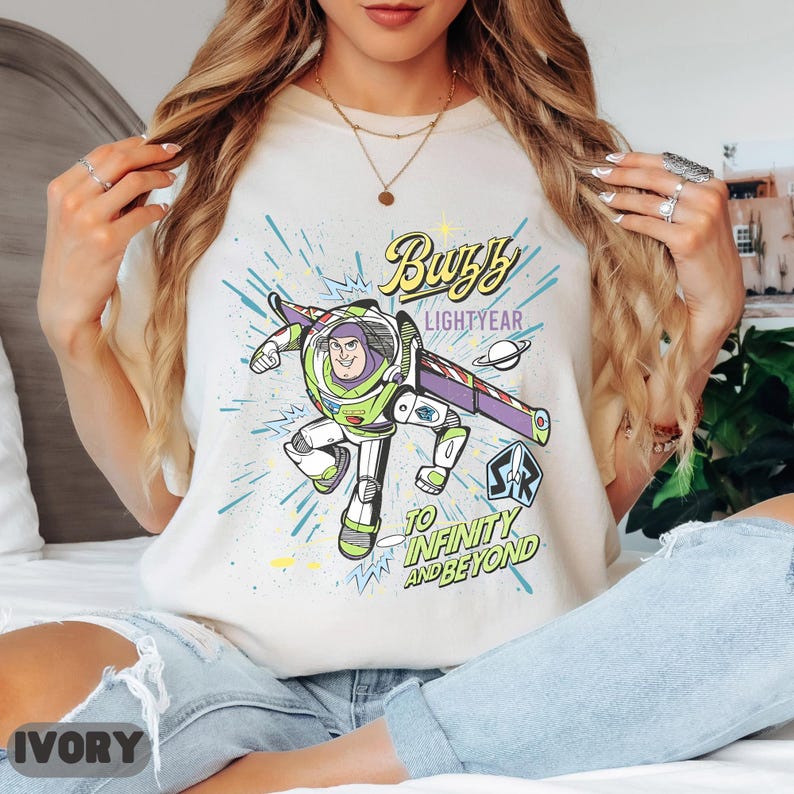 Może przedstawiać: Kremowa koszulka z grafiką Buzza Astrala w dynamicznej pozie. Projekt zawiera napisy "Buzz Lightyear" i "To Infinity and Beyond" w kolorowej czcionce w stylu komiksowym. Jest to element odzieży codziennej.