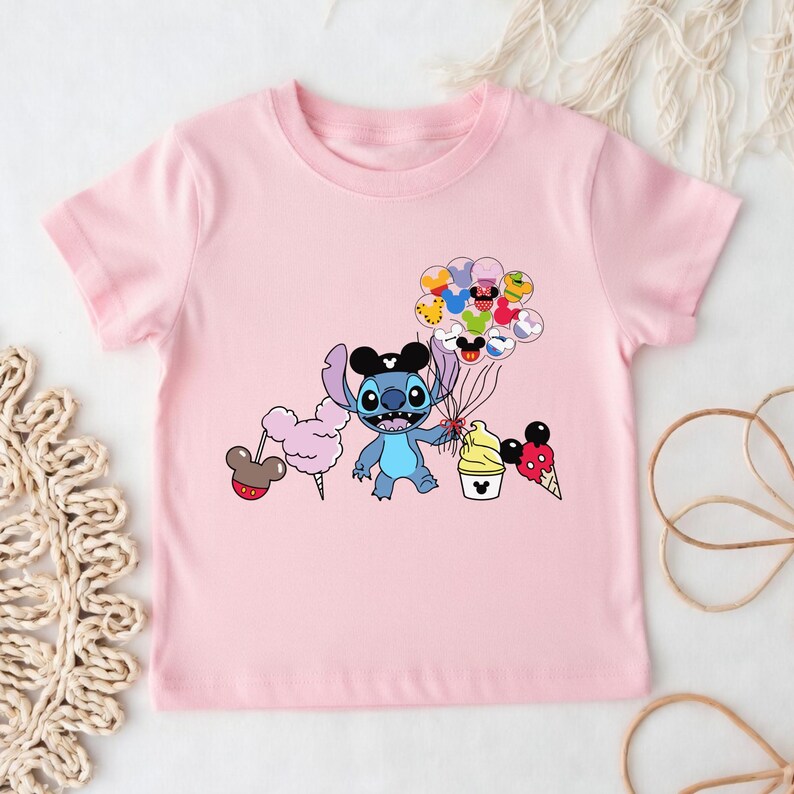 Puede incluir: Camiseta rosa de manga corta con un personaje de dibujos animados que sostiene globos, algod&oacute;n de az&uacute;car, una magdalena y un cono de helado. El personaje azul lleva un sombrero negro. Los globos tienen dise&ntilde;os de Mickey Mouse.