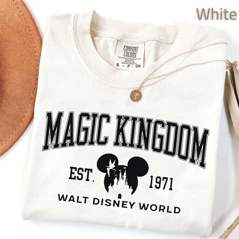 May include: White t-shirt with "MAGIC KINGDOM" in black block letters. Below the text is a black silhouette of Mickey Mouse ears with a castle and the text "EST. 1971 WALT DISNEY WORLD". A gold necklace with a pendant is draped on the shirt.