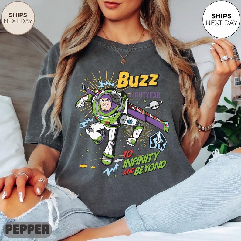 K&ouml;nnte beinhalten: Dunkelgraues T-Shirt mit einem Buzz Lightyear-Motiv und dem Text "Buzz Lightyear" und "To Infinity and Beyond". Das Design zeigt eine Cartoon-Darstellung von Buzz in seinem Raumanzug, mit einem lila-wei&szlig;en Fl&uuml;gel. Das T-Shirt hat eine lockere Passform.