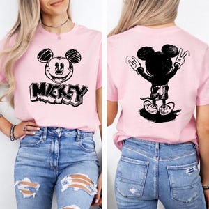 May include: A light pink t-shirt featuring a black and white graphic of Mickey Mouse on the front and back. The front design includes the word "Mickey" in a bold font. The back shows Mickey Mouse with his hands up in a rock and roll gesture.