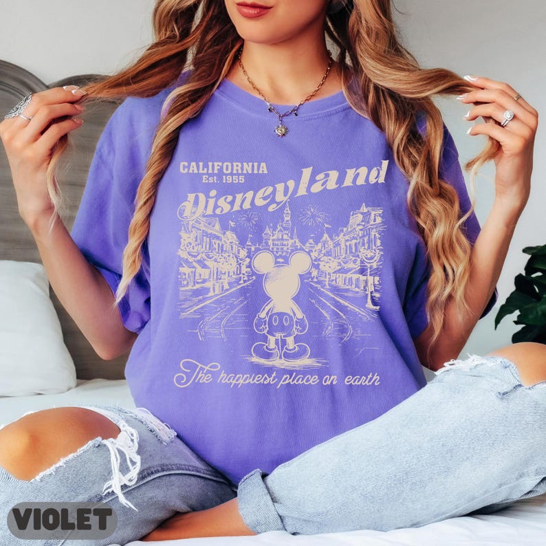 May include: Violet-colored t-shirt with a Disneyland graphic. The graphic features Mickey Mouse, the words "California Disneyland Est. 1955," and "The happiest place on earth." The shirt is a casual style.