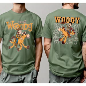 May include: Olive green t-shirt featuring a cartoon illustration of Woody riding a horse. The front of the shirt has the word "Woody" in a western-style font, with "Sheriff" above the image. The back of the shirt repeats the word "Woody" with the same illustration.