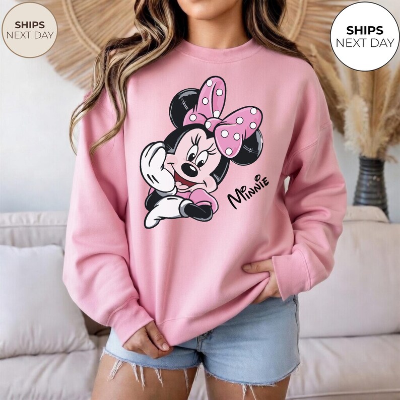 May include: A light pink sweatshirt featuring a Minnie Mouse graphic. The graphic shows Minnie Mouse with a pink polka dot bow, black ears, and the name "Minnie" in script. The sweatshirt is a crew neck style.
