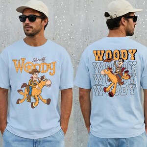 May include: Light blue t-shirt featuring a cartoon image of Woody riding a horse, with the word "Woody" in a western-style font. The back of the shirt also has a Woody graphic and the word "Woody".