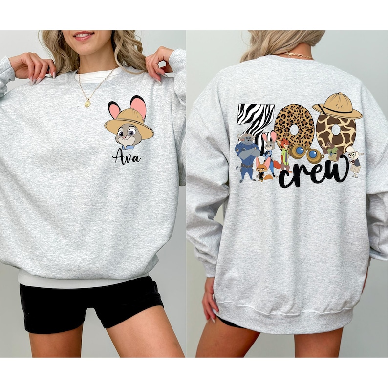 Puede incluir: Sudadera gris claro con cuello y pu&ntilde;os blancos. La parte delantera presenta un personaje de dibujos animados con un sombrero y el nombre "Ava". La parte posterior muestra "Zoo Crew" con letras y personajes tem&aacute;ticos de animales.