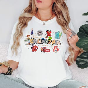 K&ouml;nnte beinhalten: Ein wei&szlig;es T-Shirt mit kurzen &Auml;rmeln und einem farbenfrohen Grafikdruck mit dem Text Disney CALIFORNIA ADVENTURE. Das Design zeigt verschiedene Zeichentrickfiguren wie Woody, Buzz Lightyear, Baymax, Lightning McQueen und Ariel.