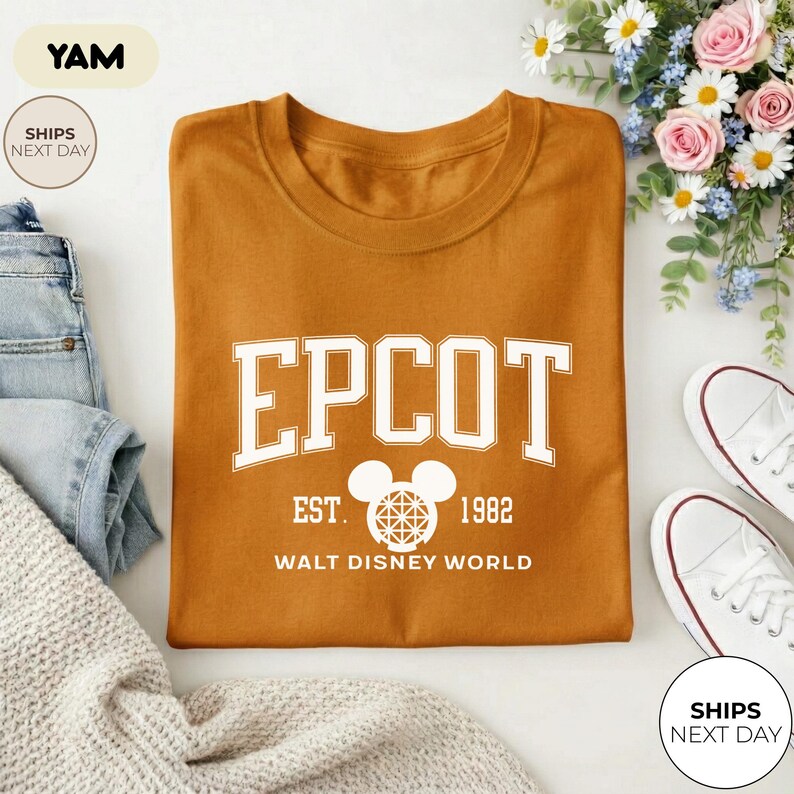 May include: Mustard-colored t-shirt with the word "EPCOT" in white block letters. Below the word "EPCOT" is "EST. 1982" and "WALT DISNEY WORLD". The shirt is folded on a white surface with a pair of white sneakers and a bouquet of flowers.