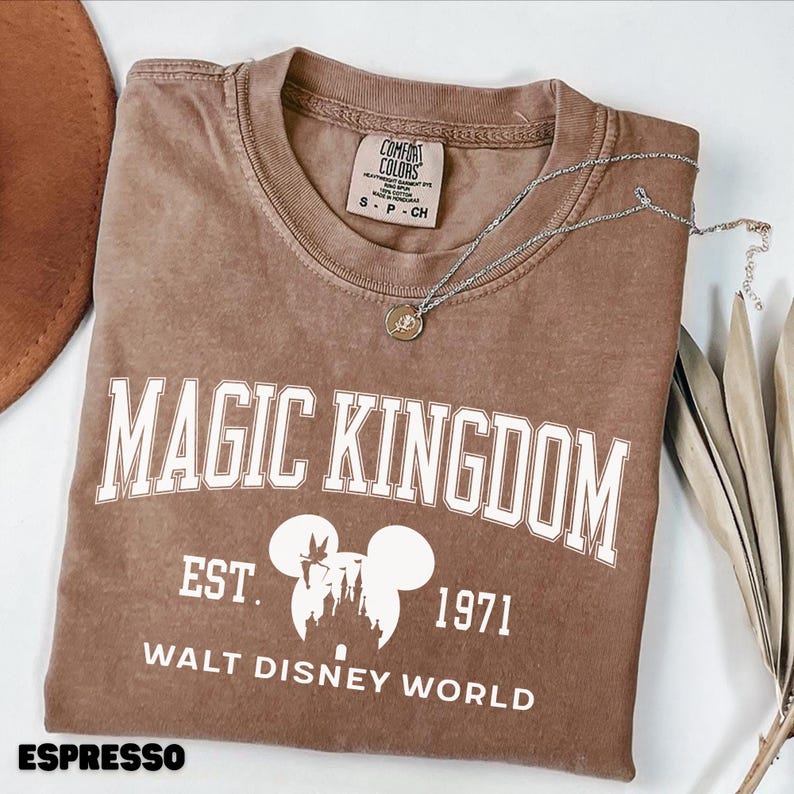 May include: Espresso-colored t-shirt with "MAGIC KINGDOM" in white block letters. Below the text is a white graphic of Mickey Mouse ears and a castle, with "EST. 1971" and "WALT DISNEY WORLD" below. A silver necklace with a pendant rests on the shirt.