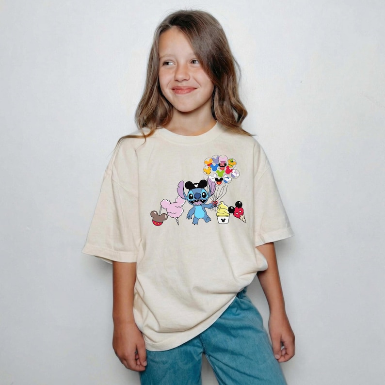 Puede incluir: Una camiseta color crema con un personaje de dibujos animados sosteniendo globos, algod&oacute;n de az&uacute;car y conos de helado. El personaje es azul con orejas grandes y lleva un sombrero negro. La camiseta la lleva una persona.