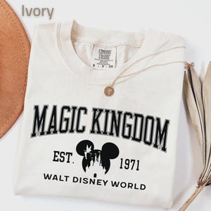 May include: Ivory-colored t-shirt with the words "MAGIC KINGDOM" in black block letters. Below the text is a silhouette of Mickey Mouse and a castle, with the text "EST. 1971 WALT DISNEY WORLD". A gold necklace and a hat are visible.