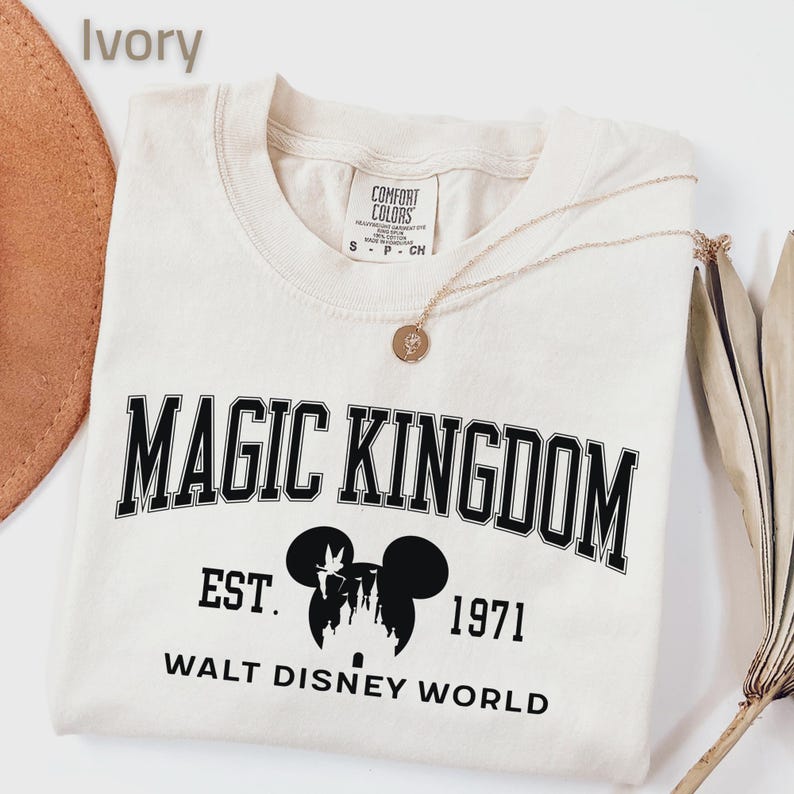 May include: Ivory-colored t-shirt with the words "MAGIC KINGDOM" in black block letters. Below the text is a silhouette of Mickey Mouse and a castle, with the text "EST. 1971 WALT DISNEY WORLD". A gold necklace and a hat are visible.