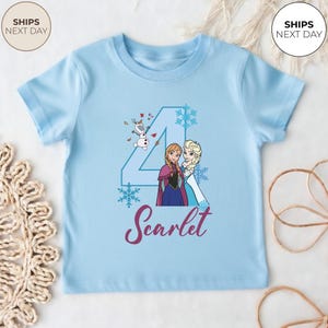 May include: A pale blue toddler's t-shirt with a Frozen-themed graphic. It features Anna and Elsa, the number 4, snowflakes, and the name "Scarlet". The design also includes Olaf.