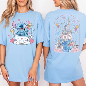 May include: Light blue t-shirt featuring a cartoon illustration of Stitch and a smaller Stitch in a teacup with hearts. The back of the shirt shows Stitch in front of a castle with fireworks and balloons.