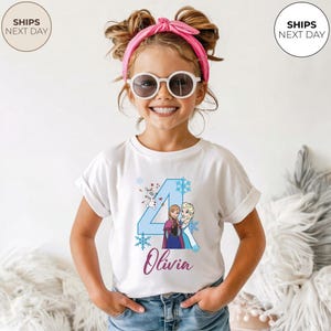 May include: A young girl wearing a white t-shirt with a blue number 4 graphic featuring characters from the movie Frozen. The shirt also has the name "Olivia" in a cursive font. She is wearing sunglasses and a pink headband.
