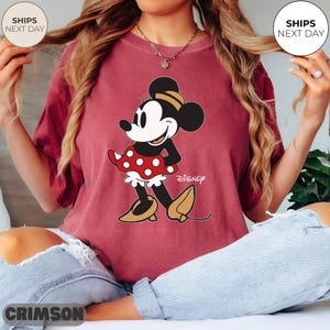 Puede incluir: Camiseta de color carmes&iacute; con un dise&ntilde;o de Minnie Mouse. El personaje de dibujos animados lleva un vestido rojo de lunares y zapatos amarillos. El logotipo de Disney est&aacute; impreso debajo. Cuello redondo y mangas cortas.
