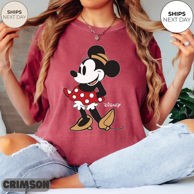 Puede incluir: Camiseta de color carmes&iacute; con un dise&ntilde;o de Minnie Mouse. El personaje de dibujos animados lleva un vestido rojo de lunares y zapatos amarillos. El logotipo de Disney est&aacute; impreso debajo. Cuello redondo y mangas cortas.