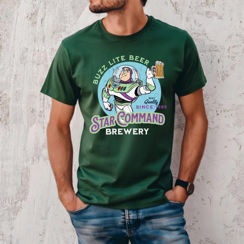 May include: A dark green short-sleeve t-shirt featuring a graphic of a space ranger character holding a beer mug. The text reads BUZZ LITE BEER, STAR COMMAND BREWERY, and BEST QUALITY SINCE 1995. The design is set against a light blue circular background.