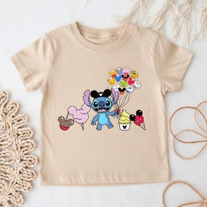 Puede incluir: Camiseta beige con una ilustraci&oacute;n de dibujos animados de Stitch sosteniendo globos y varias golosinas. El dise&ntilde;o incluye algod&oacute;n de az&uacute;car, un cono de helado y una taza de helado. Los globos tienen caras de Mickey Mouse.