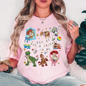 May include: Pale pink t-shirt with a colourful cartoon design. The design features characters from a popular animated film, including Woody and Buzz Lightyear, and the text "YOU'VE GOT A FRIEND IN ME."
