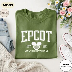 May include: An olive green t-shirt with the word "EPCOT" in white block letters, along with "EST. 1982" and "WALT DISNEY WORLD". The shirt is folded and surrounded by jeans, a sweater, and white sneakers.