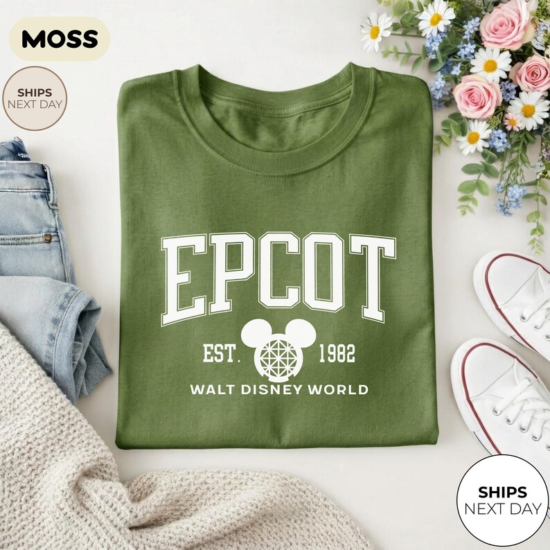 May include: An olive green t-shirt with the word "EPCOT" in white block letters, along with "EST. 1982" and "WALT DISNEY WORLD". The shirt is folded and surrounded by jeans, a sweater, and white sneakers.