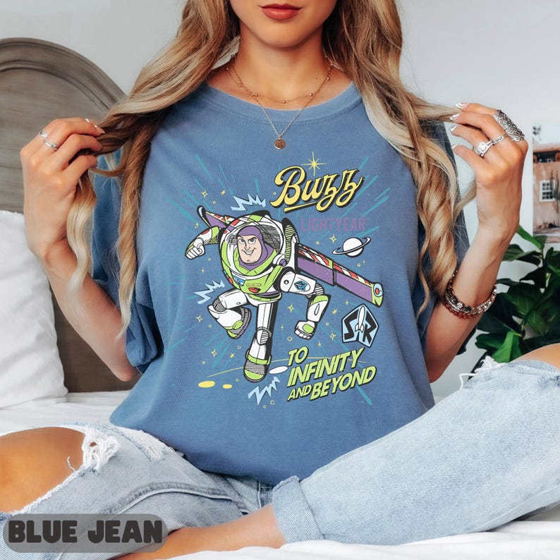 Może przedstawiać: Niebieska koszulka z grafiką Buzza Astrala, napisem "Buzz Lightyear" i "To Infinity and Beyond". Projekt zawiera kresk&oacute;wkowy wizerunek Buzza w jego skafandrze kosmicznym, z fioletowo-białym statkiem kosmicznym. Koszulka jest w jednolitym niebieskim kolorze.