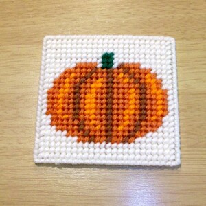May include: A white canvas with a brown, orange, and green pumpkin design. The pumpkin is stitched with yarn.