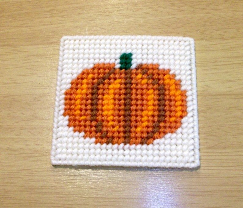 May include: A white canvas with a brown, orange, and green pumpkin design. The pumpkin is stitched with yarn.
