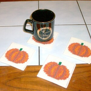 May include: Four white crocheted coasters with a pumpkin design in orange and brown. A black coffee mug with a gold logo that says "St. Louis Cardinals" is sitting on one of the coasters.