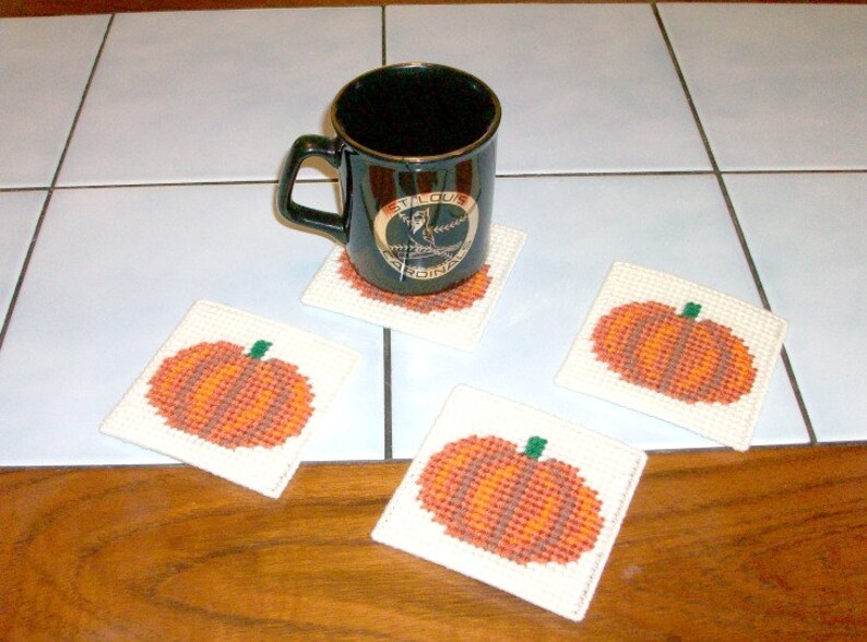 May include: Four white crocheted coasters with a pumpkin design in orange and brown. A black coffee mug with a gold logo that says "St. Louis Cardinals" is sitting on one of the coasters.