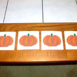May include: Four white coasters with embroidered orange pumpkins on a wooden surface.