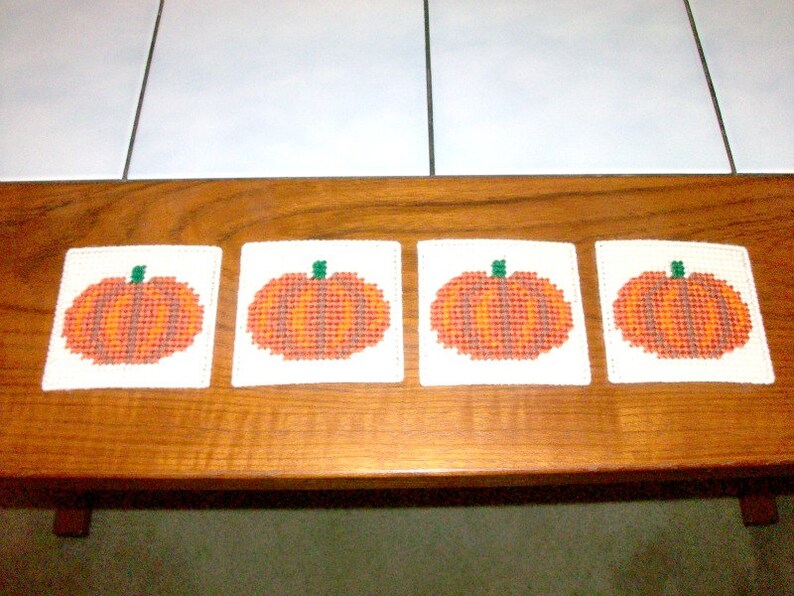 May include: Four white coasters with embroidered orange pumpkins on a wooden surface.