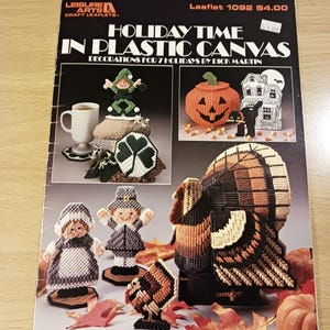 May include: A Leisure Arts craft leaflet titled Holiday Time in Plastic Canvas by Dick Martin, featuring various holiday-themed plastic canvas projects including a leprechaun, pumpkin, haunted house, black cat, turkey, and pilgrim figures.