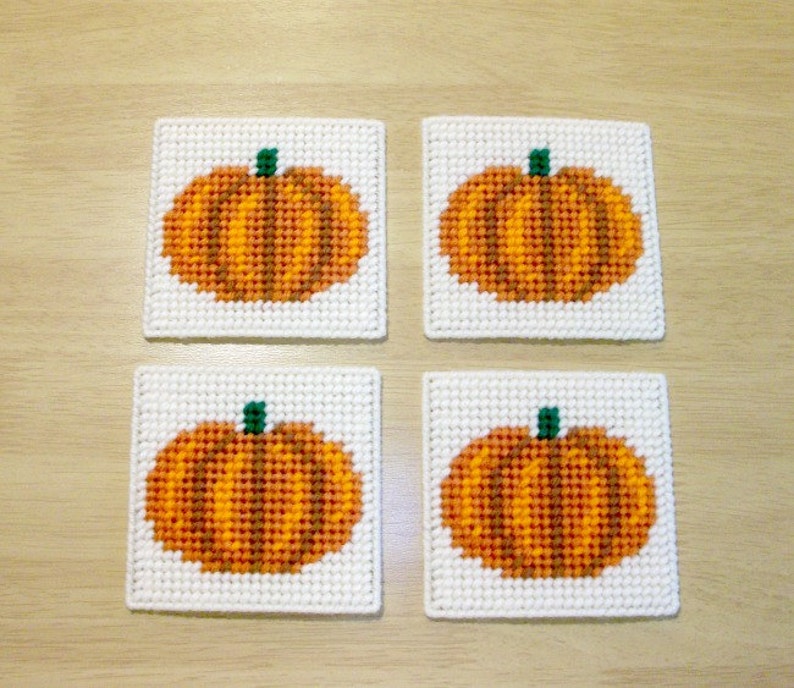 May include: Four white square coasters with a stitched design of orange pumpkins with green stems.