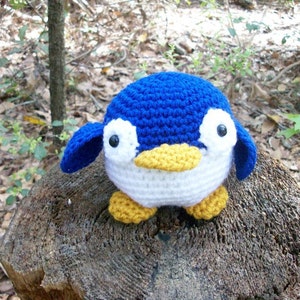 May include: A crocheted penguin toy with blue and white body, yellow beak and feet. The penguin is sitting on a tree stump.